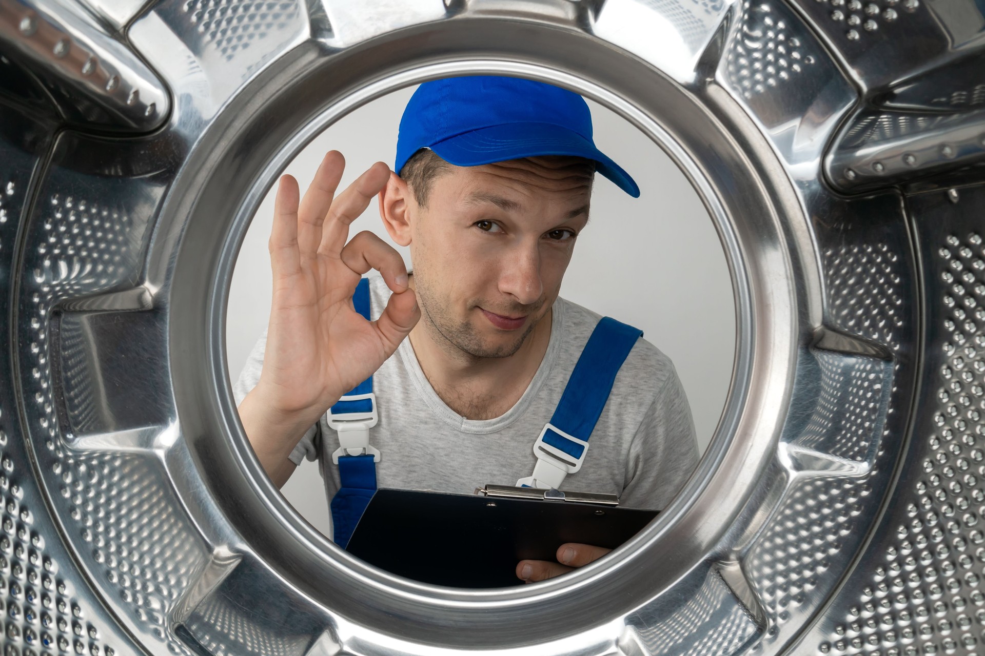 Un réparateur en uniforme avec une tablette à la main montre le signe OK, regarde dans le tambour de la machine à laver, photo de l’intérieur.